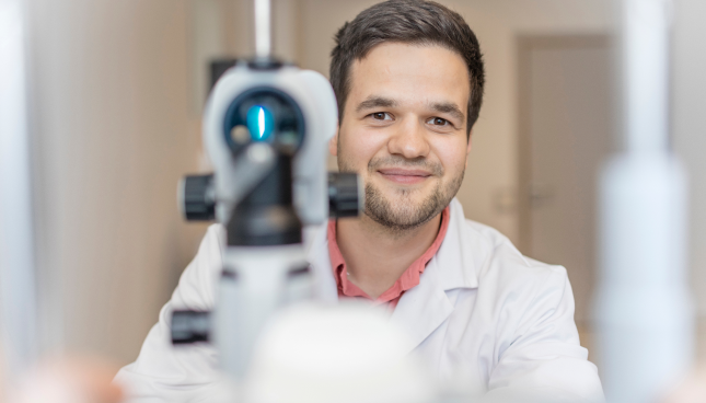 Friendly looking male optometrist standing behind eye exam  equipment used to fit contact lenses